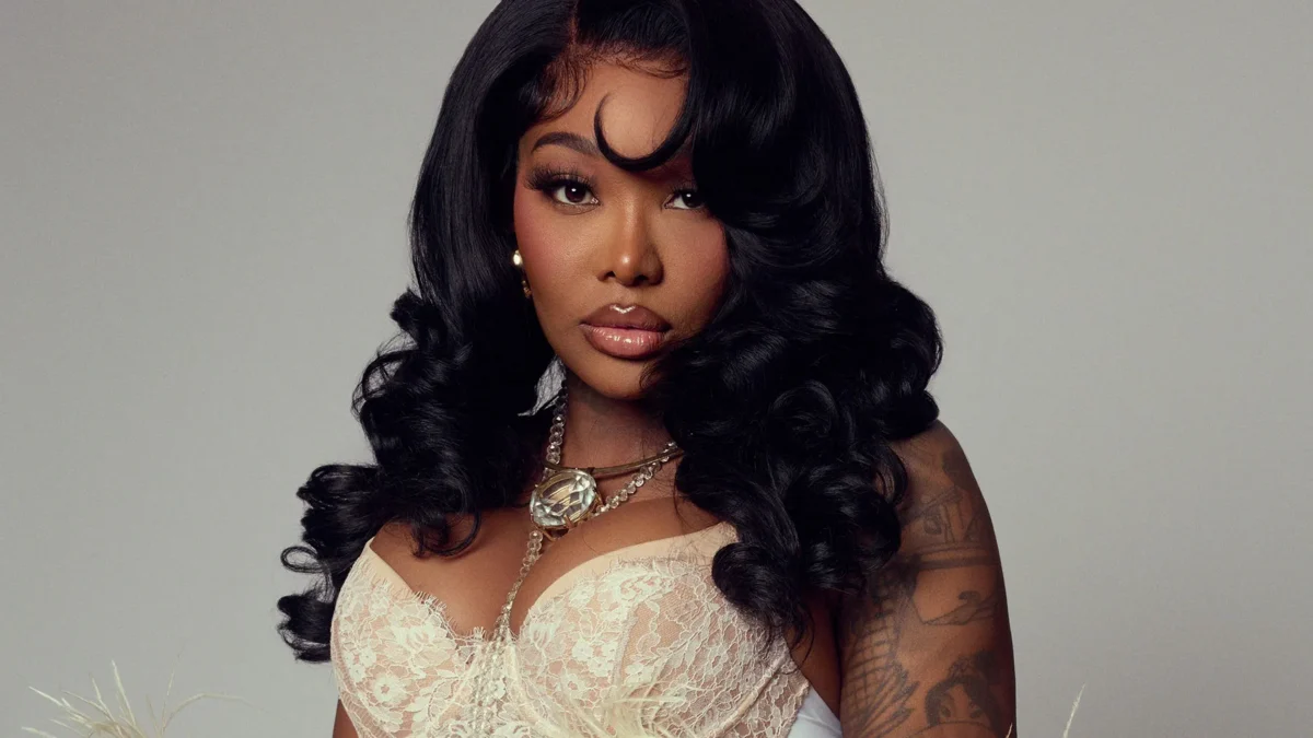 A woman with long black curls wears a white lace top, jewelry, and a feathered wrap against a gray background.