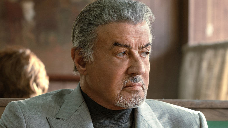 Older man with gray hair and a goatee looks sideways while seated indoors in a light blazer.