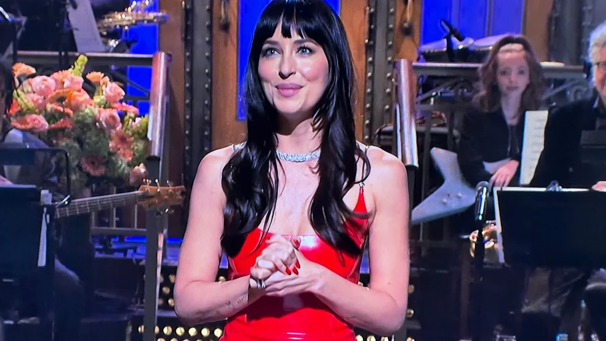A woman in a red dress stands on a stage with musicians and flowers behind her.