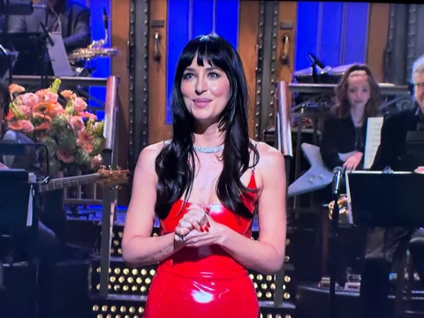 A woman in a red dress stands on a stage with musicians and flowers behind her.