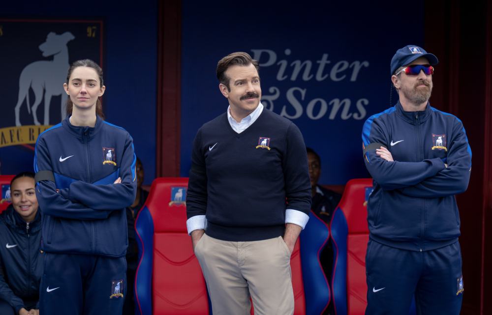 Three sports staff members stand in front of red stadium seats, wearing navy team gear.