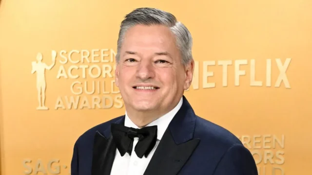 A smiling gray-haired man in a tuxedo poses before a gold backdrop with awards and Netflix lettering.