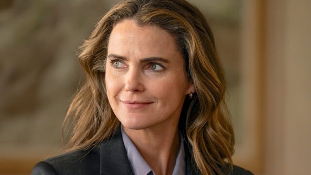 A woman with wavy brown hair in a dark blazer smiles slightly while looking to the side.