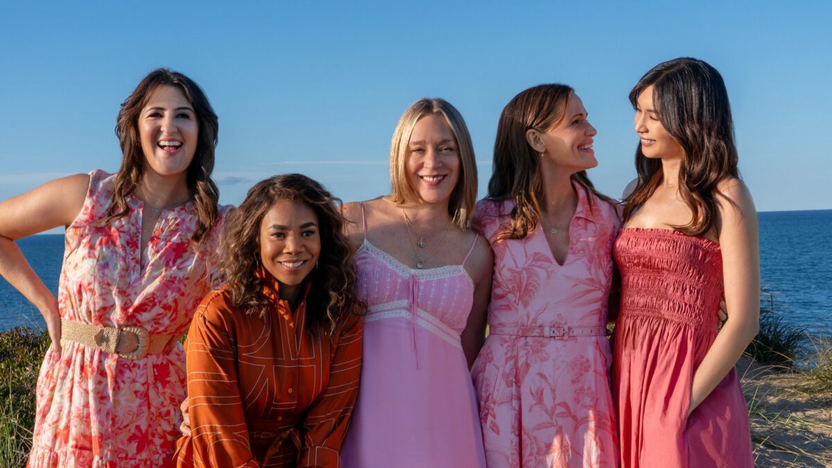 Five women in colorful dresses smile together outdoors with the ocean and blue sky behind them.