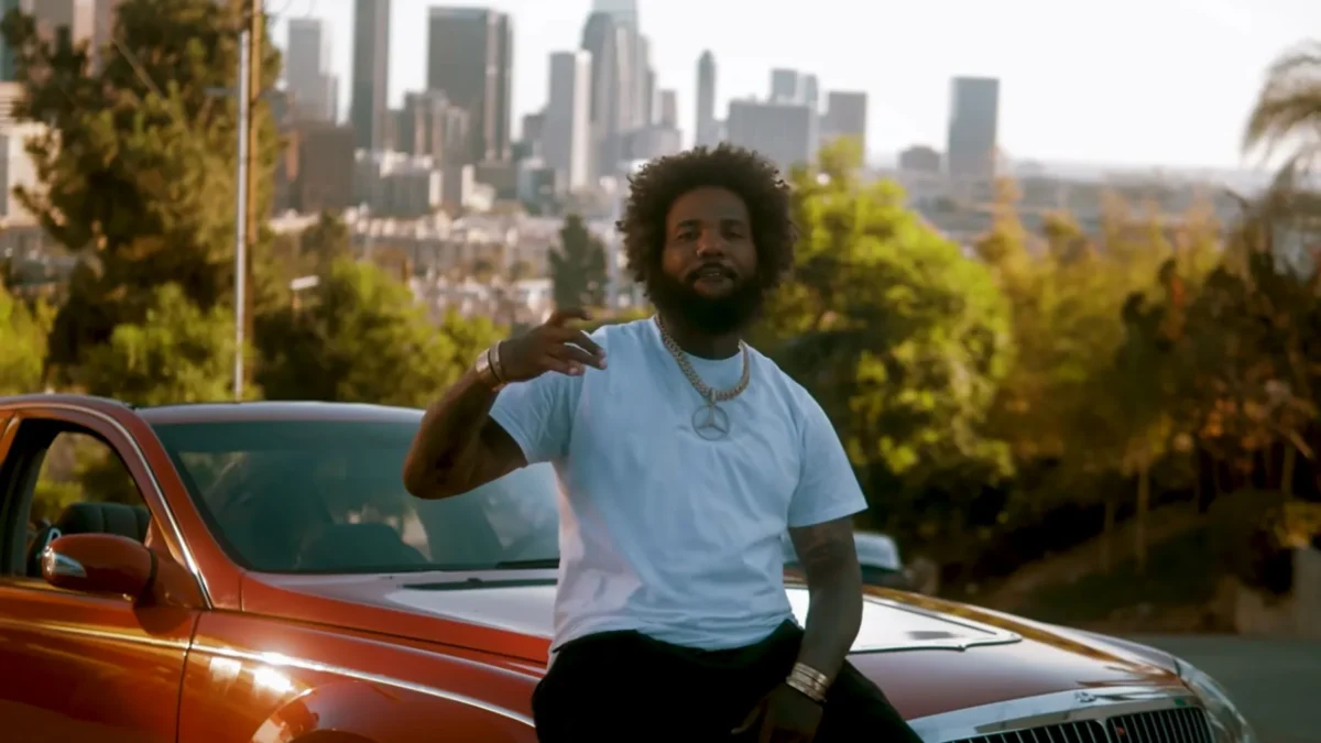 A man in a white T-shirt sits on an orange car with a blurred city skyline behind him.