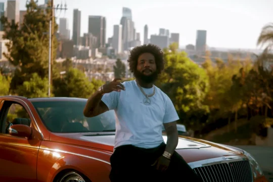 A man in a white T-shirt sits on an orange car with a blurred city skyline behind him.