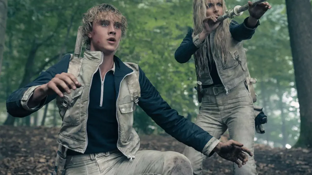 Two mud-streaked people in beige gear brace in a forest, one aiming a metal tube.