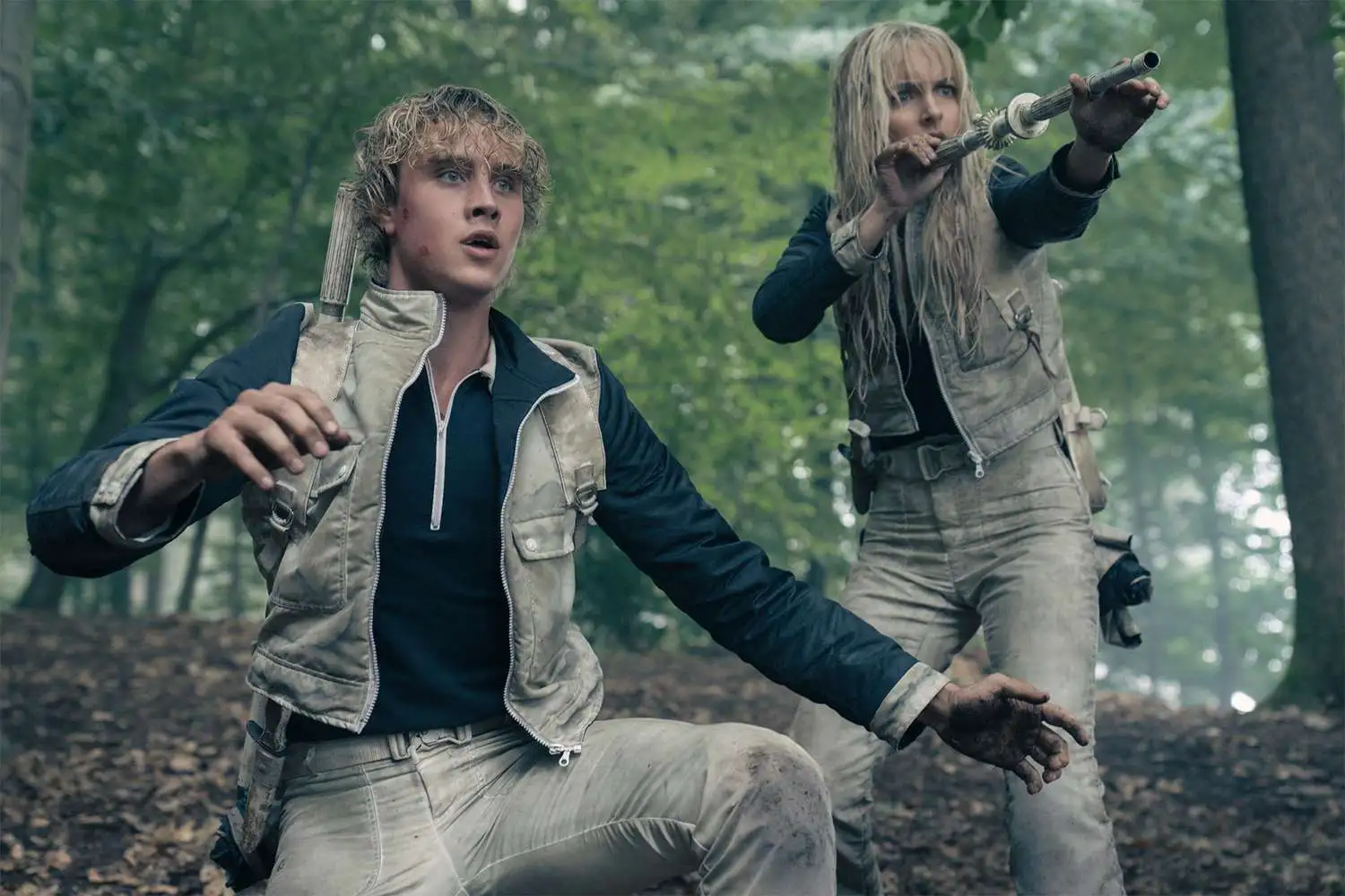 Two mud-streaked people in beige gear brace in a forest, one aiming a metal tube.