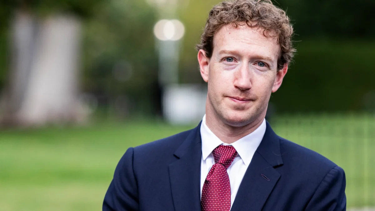 A man in a navy suit and red tie stands before a blurred green outdoor background.