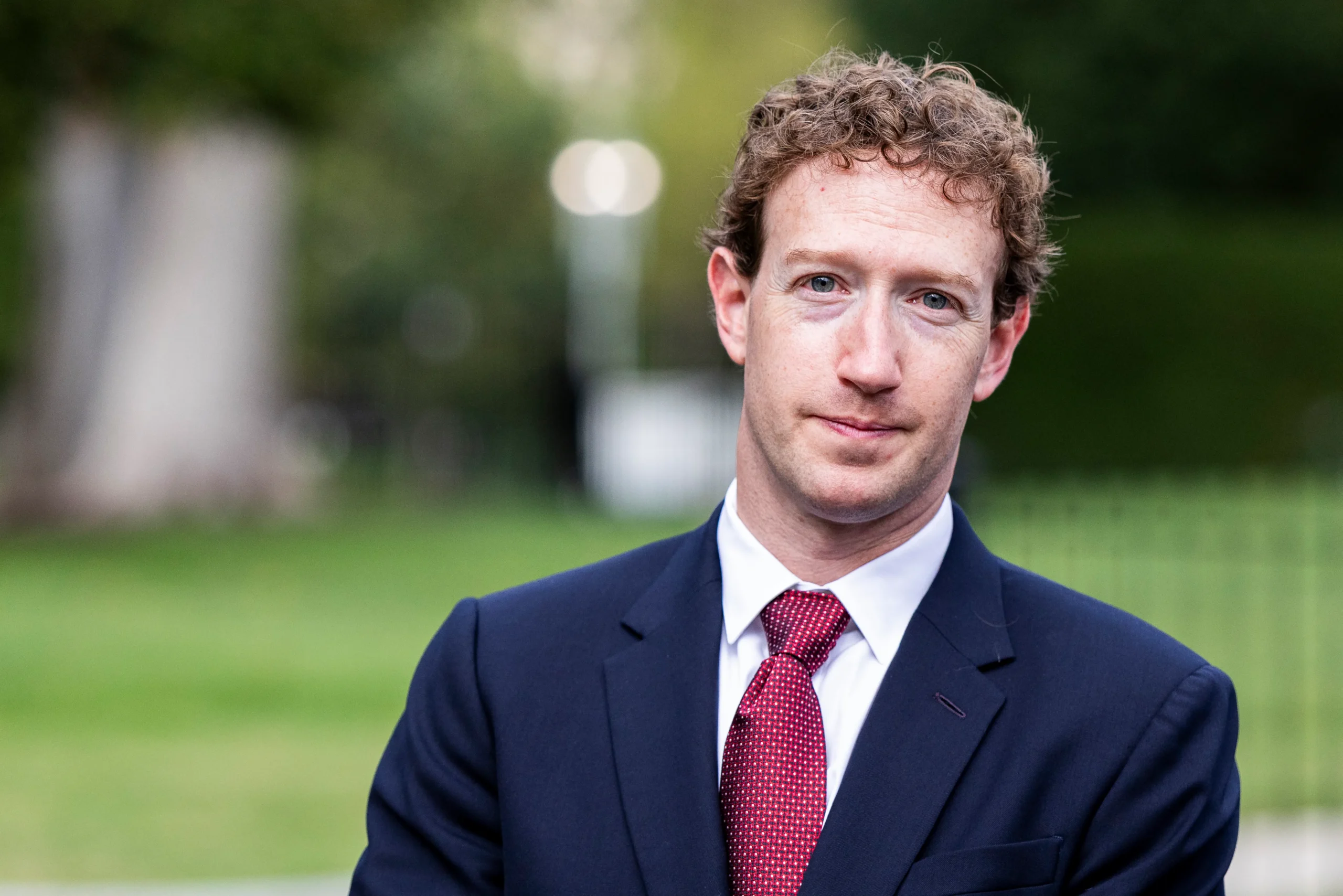 A man in a navy suit and red tie stands before a blurred green outdoor background.