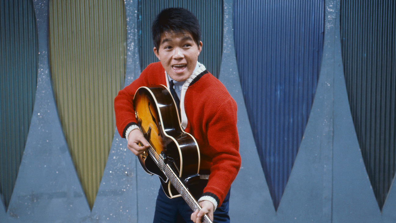 A young man in a red sweater strums an electric guitar against a colorful pleated backdrop.