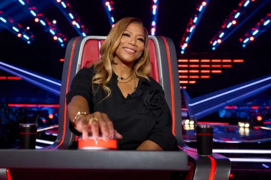 A smiling woman seated in a red chair presses a large button amid colorful stage lights.