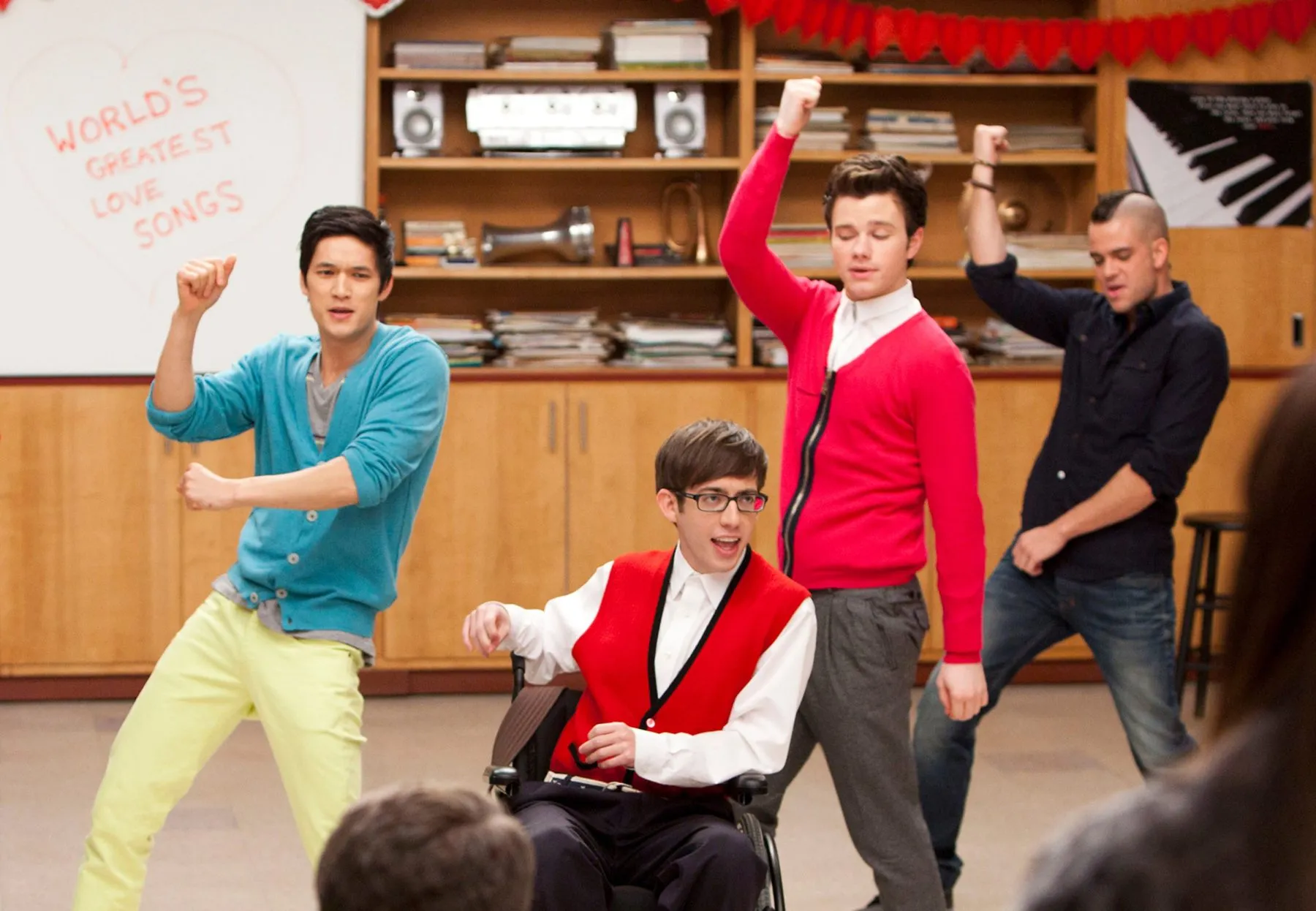 Four people dance in a room, one seated in a wheelchair, with shelves and a whiteboard behind them.