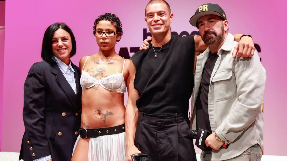Four people pose together onstage against a pink backdrop, with two holding microphones.