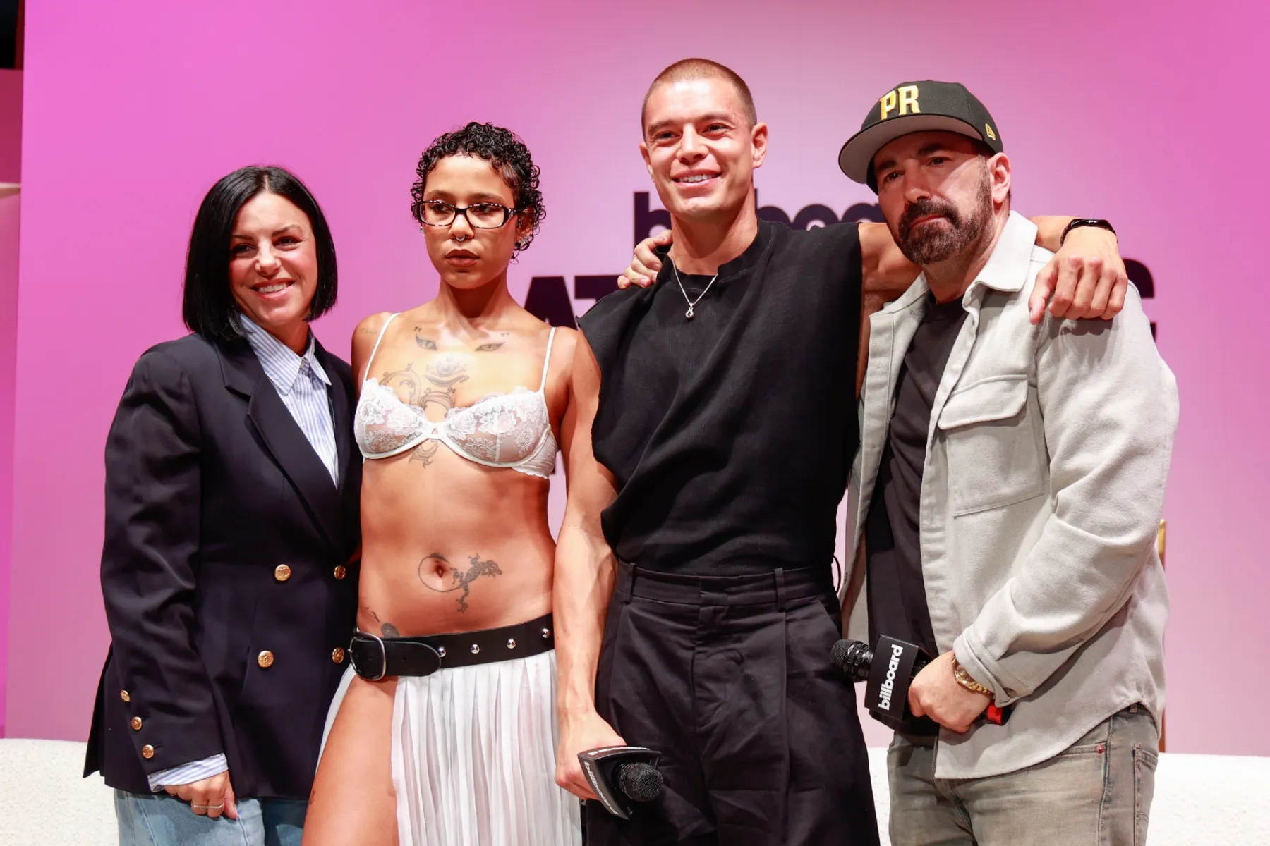 Four people pose together onstage against a pink backdrop, with two holding microphones.