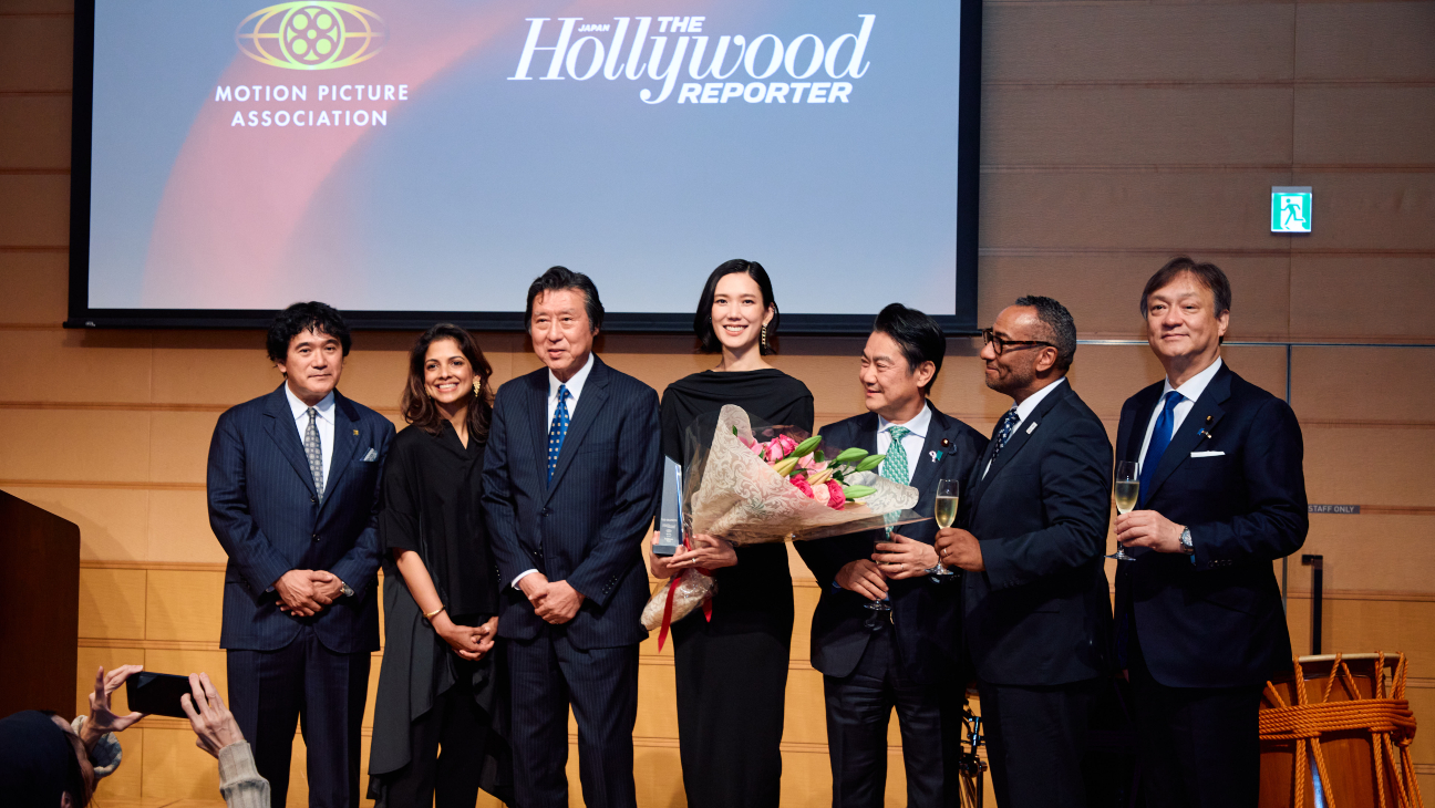 Seven formally dressed people stand onstage, one holding flowers and a clear plaque beneath large presentation screen logos.