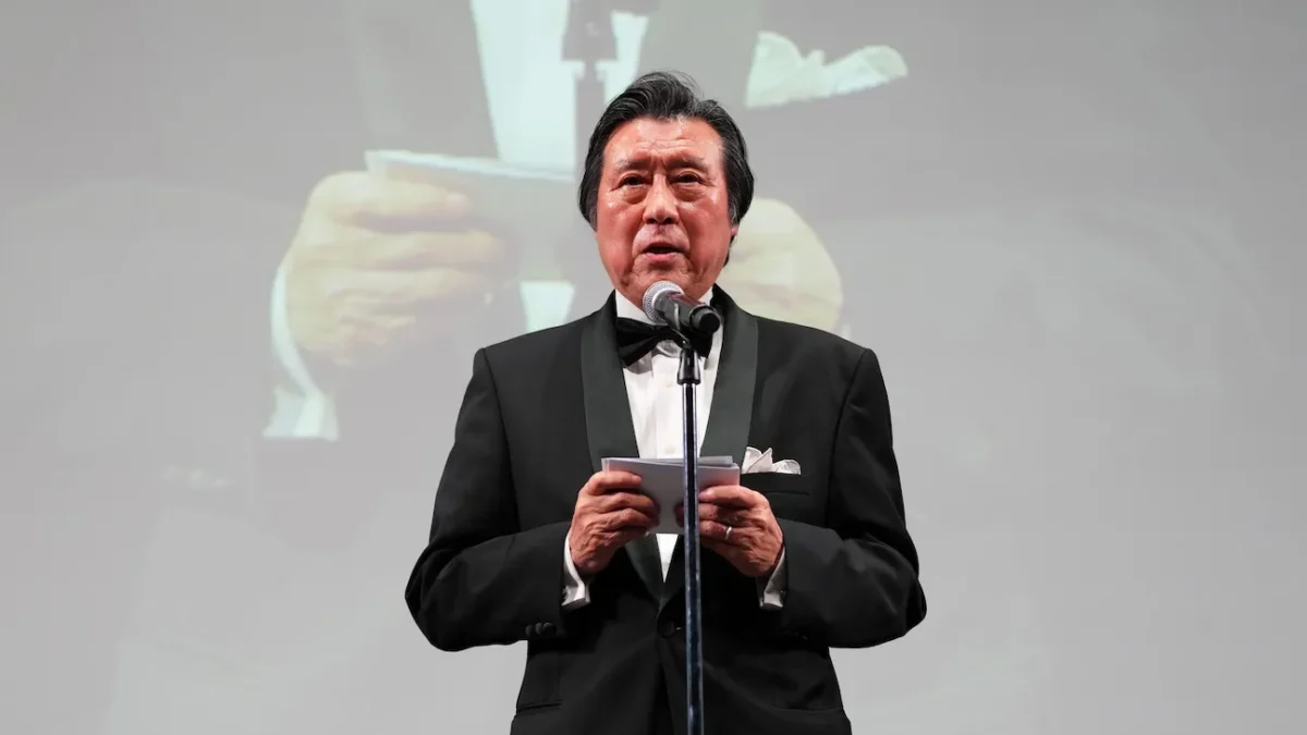 A man in a black tuxedo speaks at a microphone while holding notes on a stage.