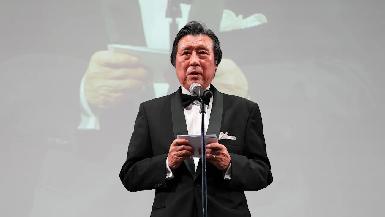 A man in a black tuxedo speaks at a microphone while holding notes on a stage.