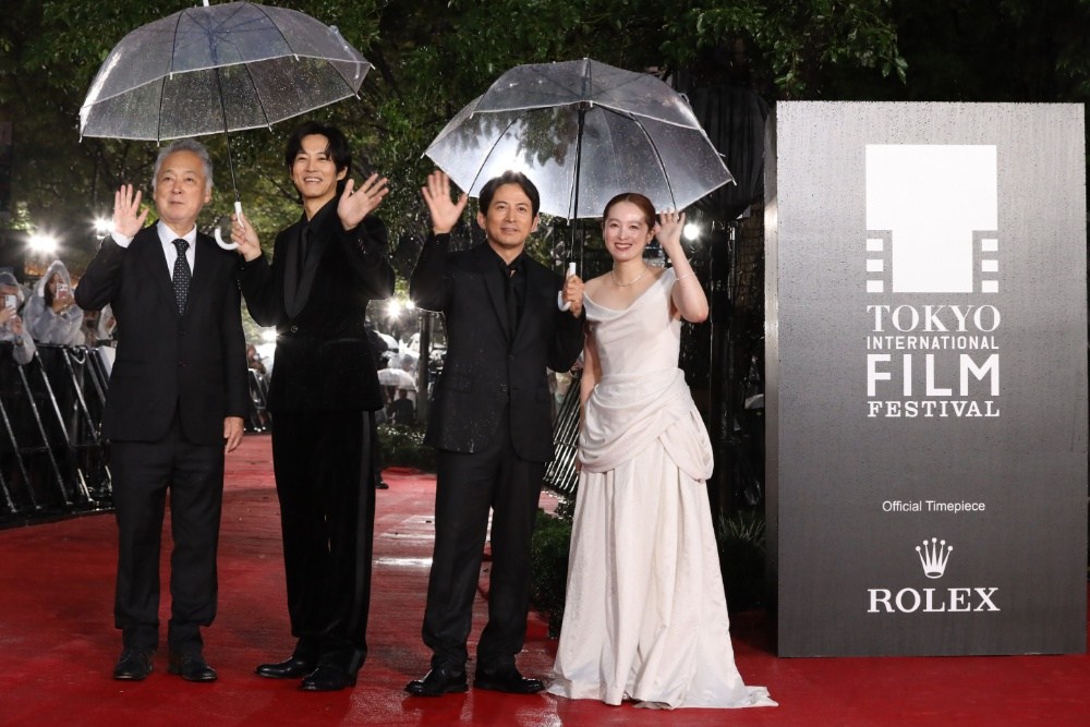 Four formally dressed people wave under clear umbrellas on a rainy red carpet beside a film festival sign.