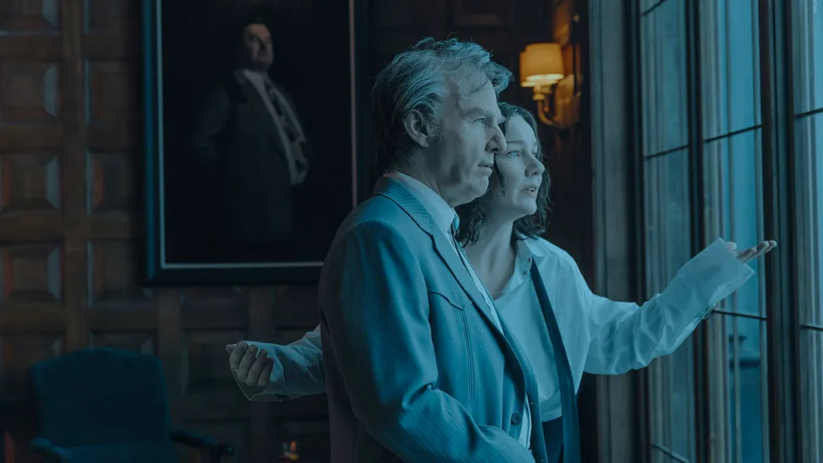 An older man and a woman stand by tall windows in a dim room with a portrait behind them.