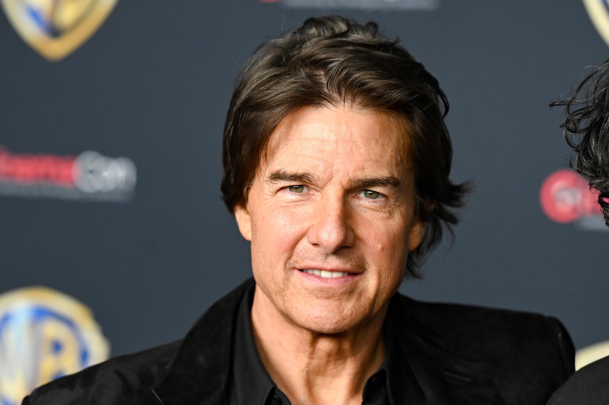 A dark-haired man in a black jacket smiles in front of a dark backdrop with blurred logos.