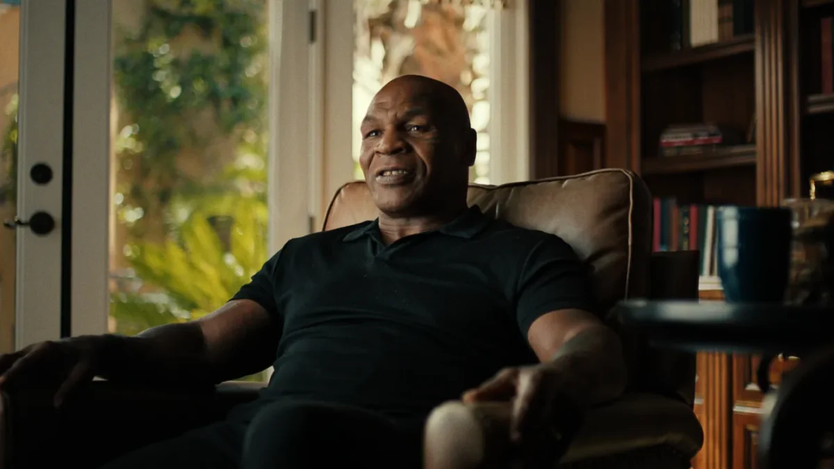 A bald man in a black polo sits in an armchair beside bookshelves and sunlit windows.