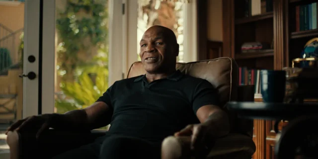 A bald man in a black polo sits in an armchair beside bookshelves and sunlit windows.