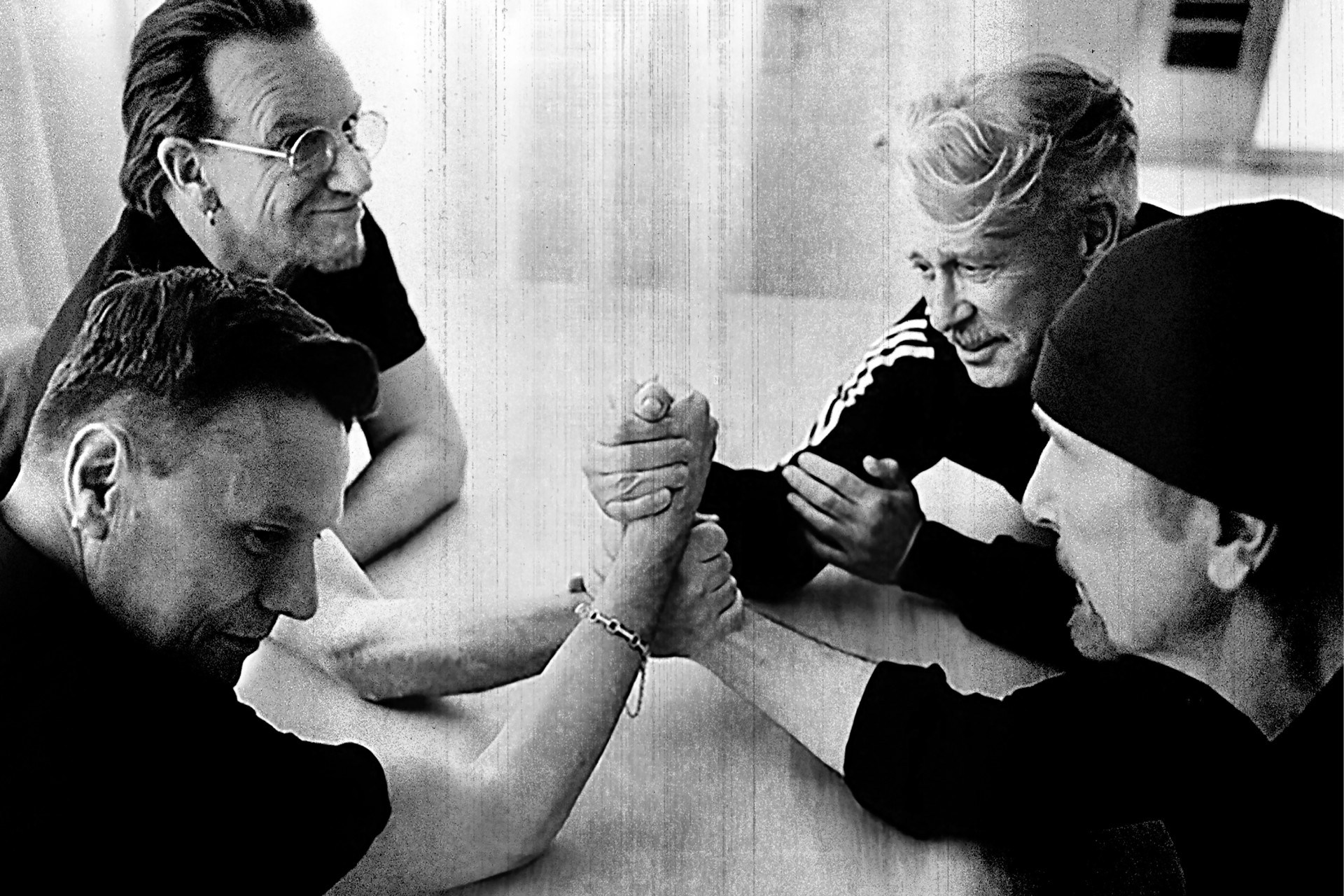 Four men gather around a table as two clasp hands in a black-and-white, grainy scene.