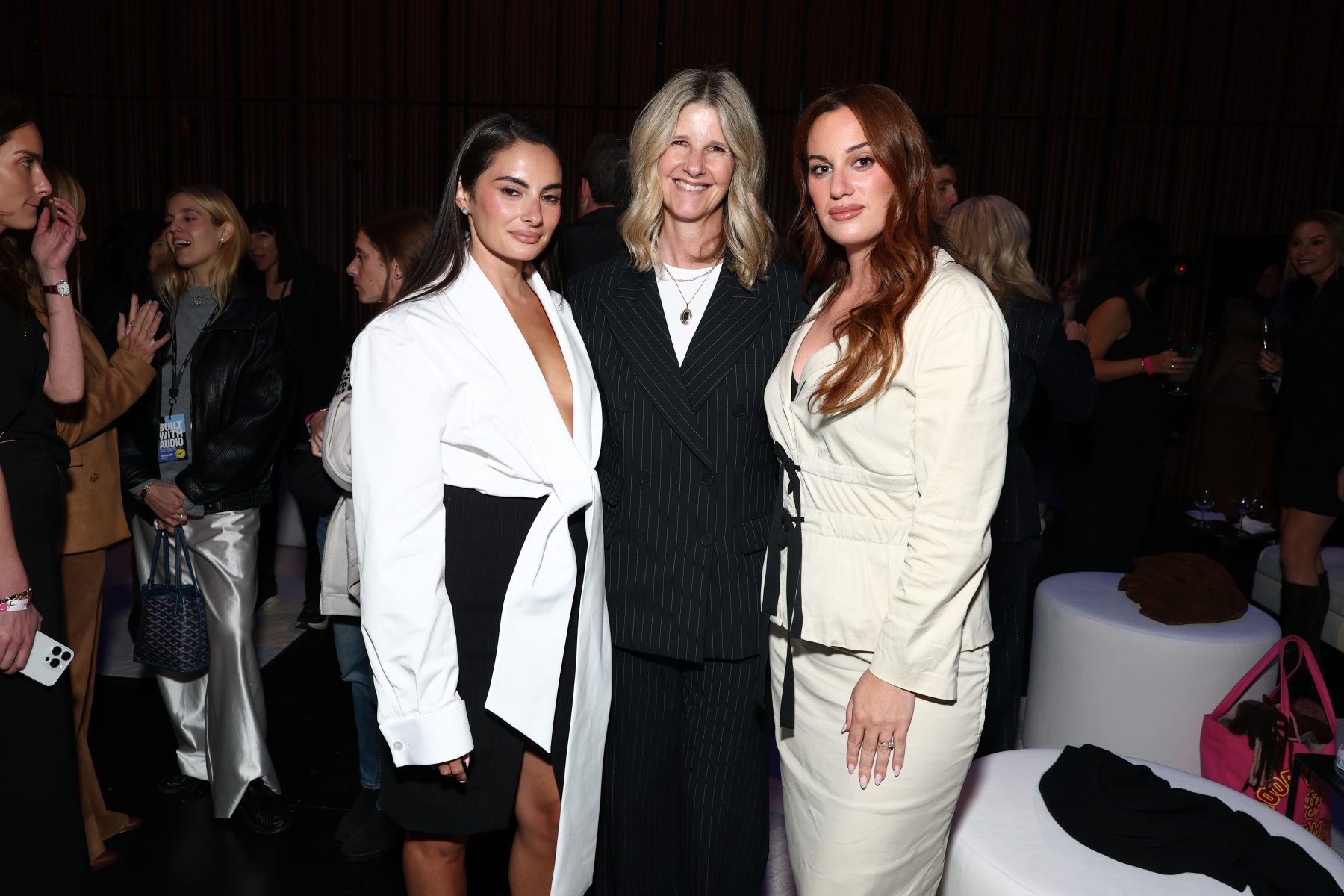 Three women in suits pose together at a crowded indoor event.