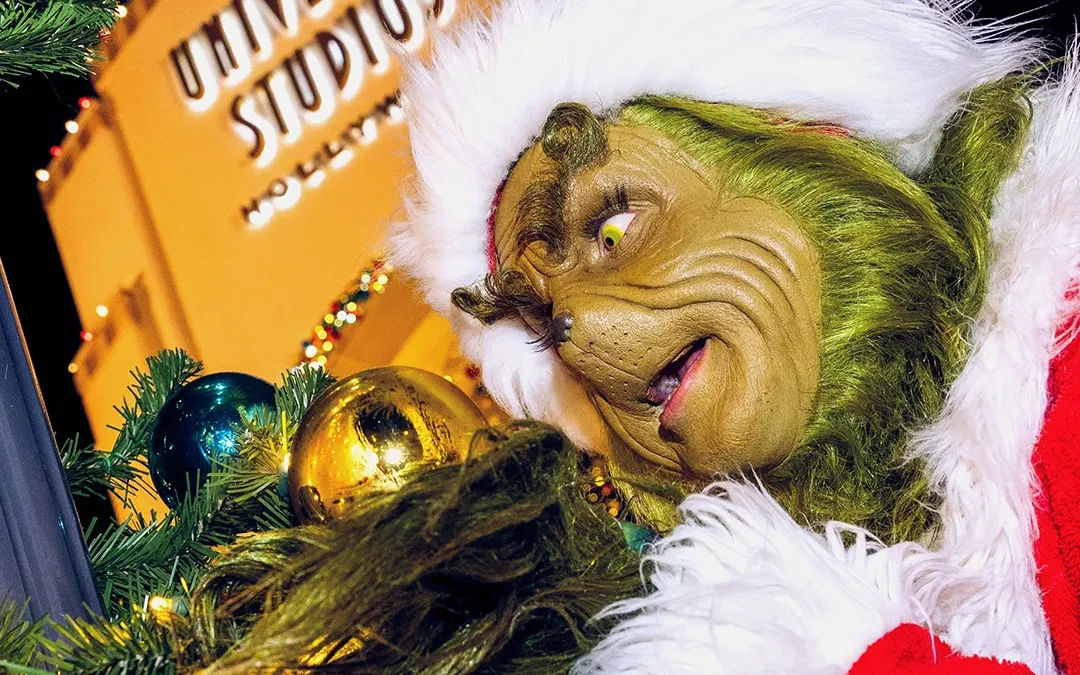 A green furry character in a red Santa outfit peers at ornaments beside a lit Universal Studios sign.