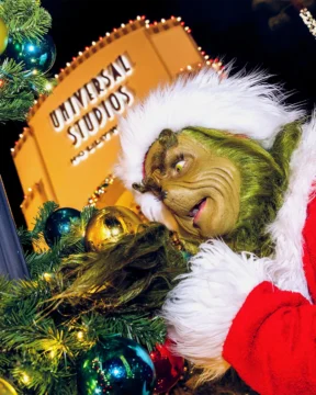 A green furry character in a red Santa outfit peers at ornaments beside a lit Universal Studios sign.