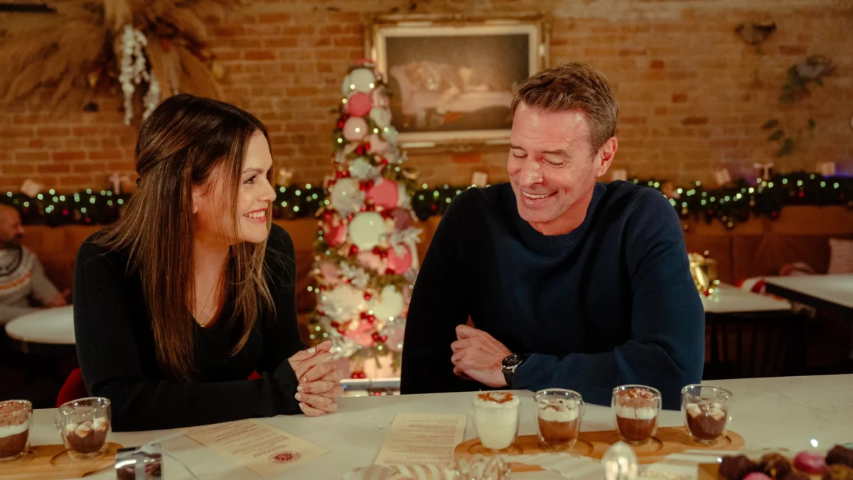 Two smiling adults sit at a dessert counter in a warmly lit room with a decorated tree.