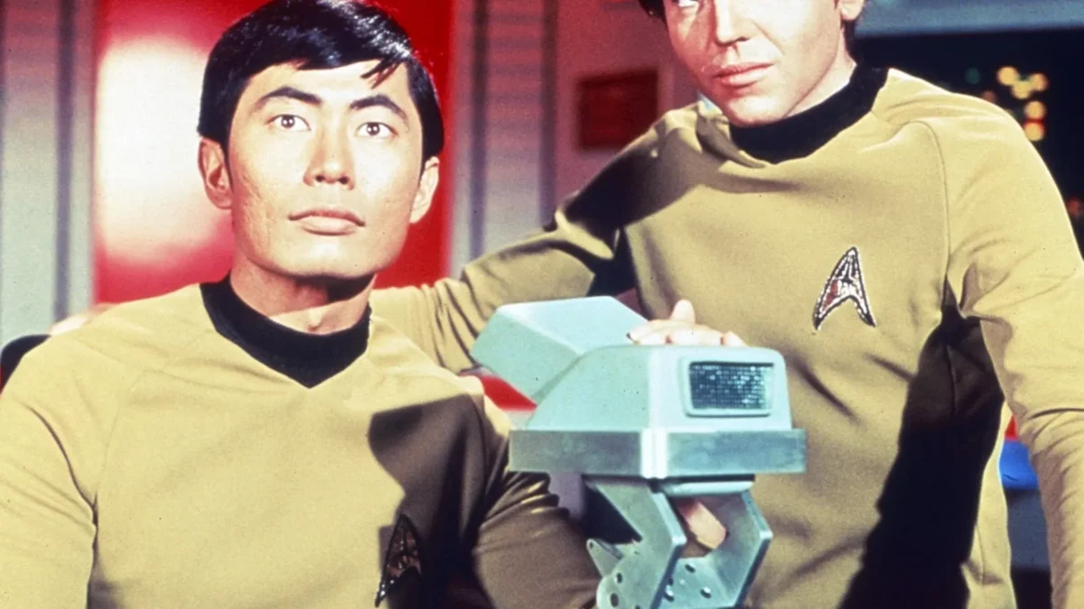 Two men in gold uniforms sit at a control console on a futuristic set.