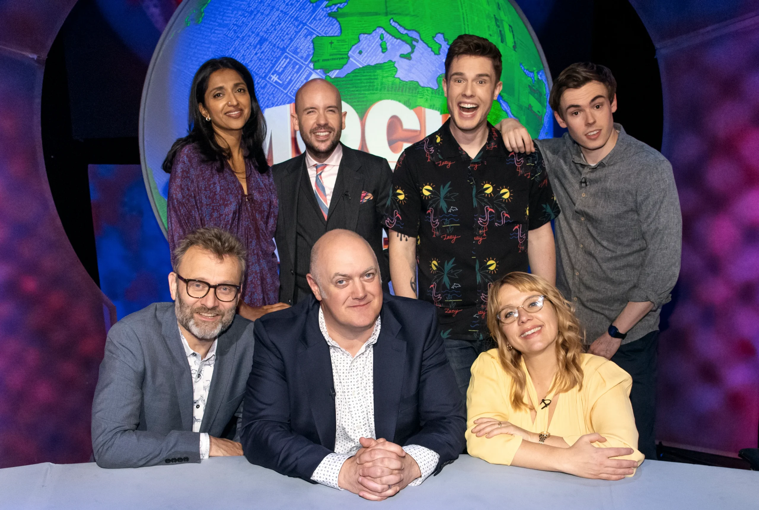 Seven people pose in a studio before a large colorful globe backdrop.