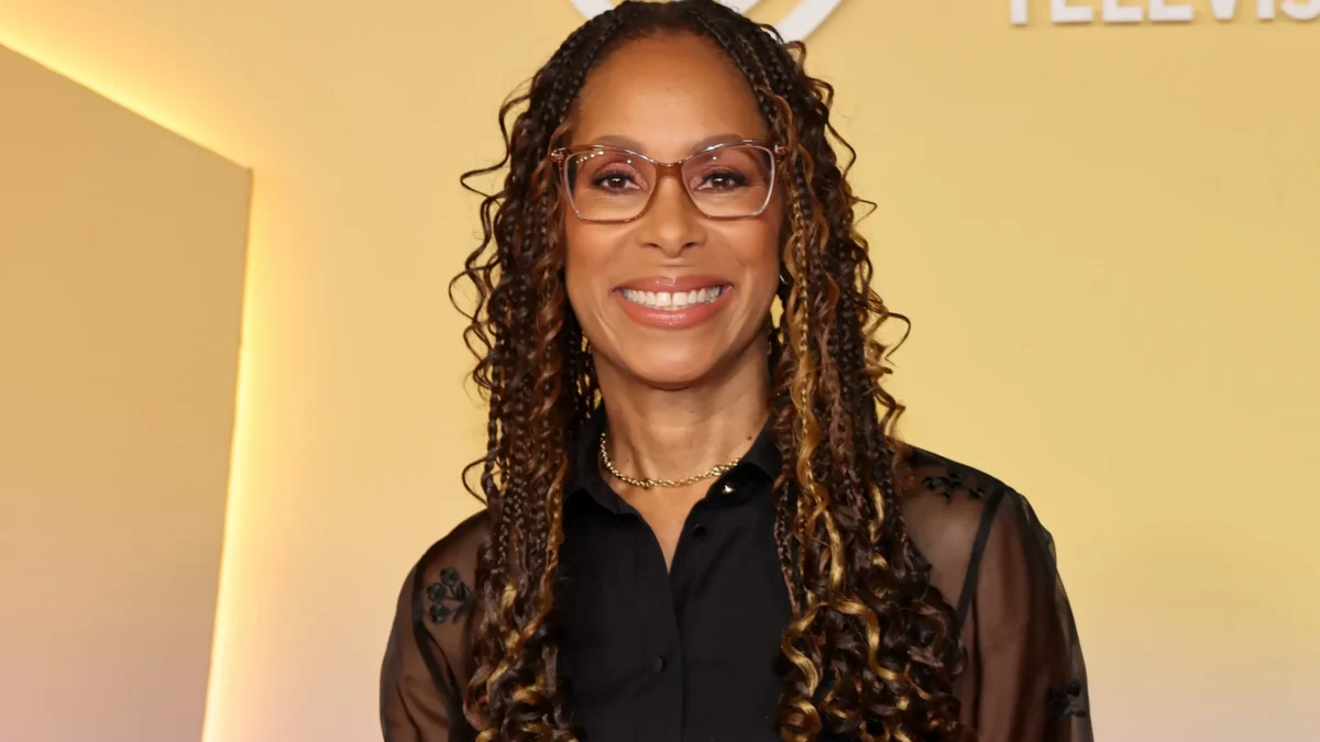 A smiling woman with glasses and long highlighted braids wears a black sheer-sleeved blouse against a yellow backdrop.