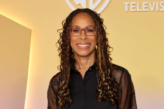 A smiling woman with glasses and long highlighted braids wears a black sheer-sleeved blouse against a yellow backdrop.