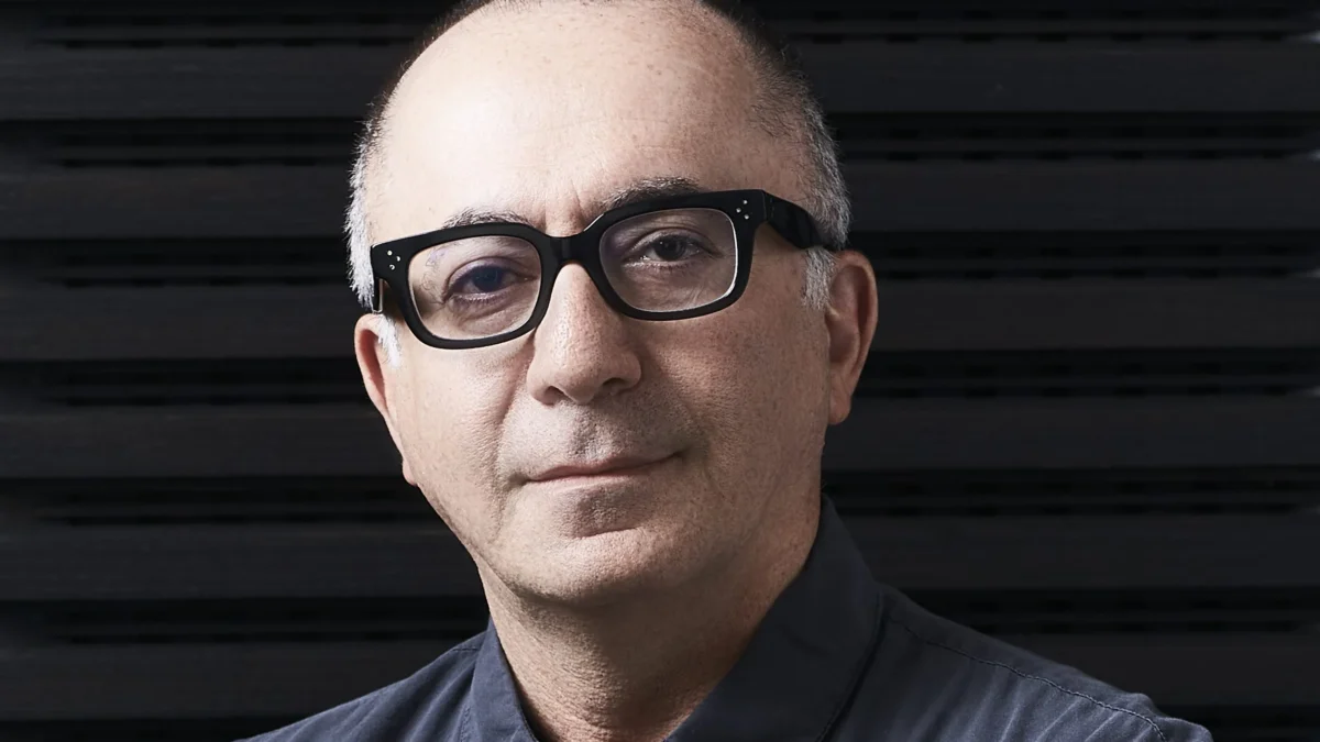 Close-up portrait of a bald man wearing black glasses and a dark button-up shirt against a black slatted background.