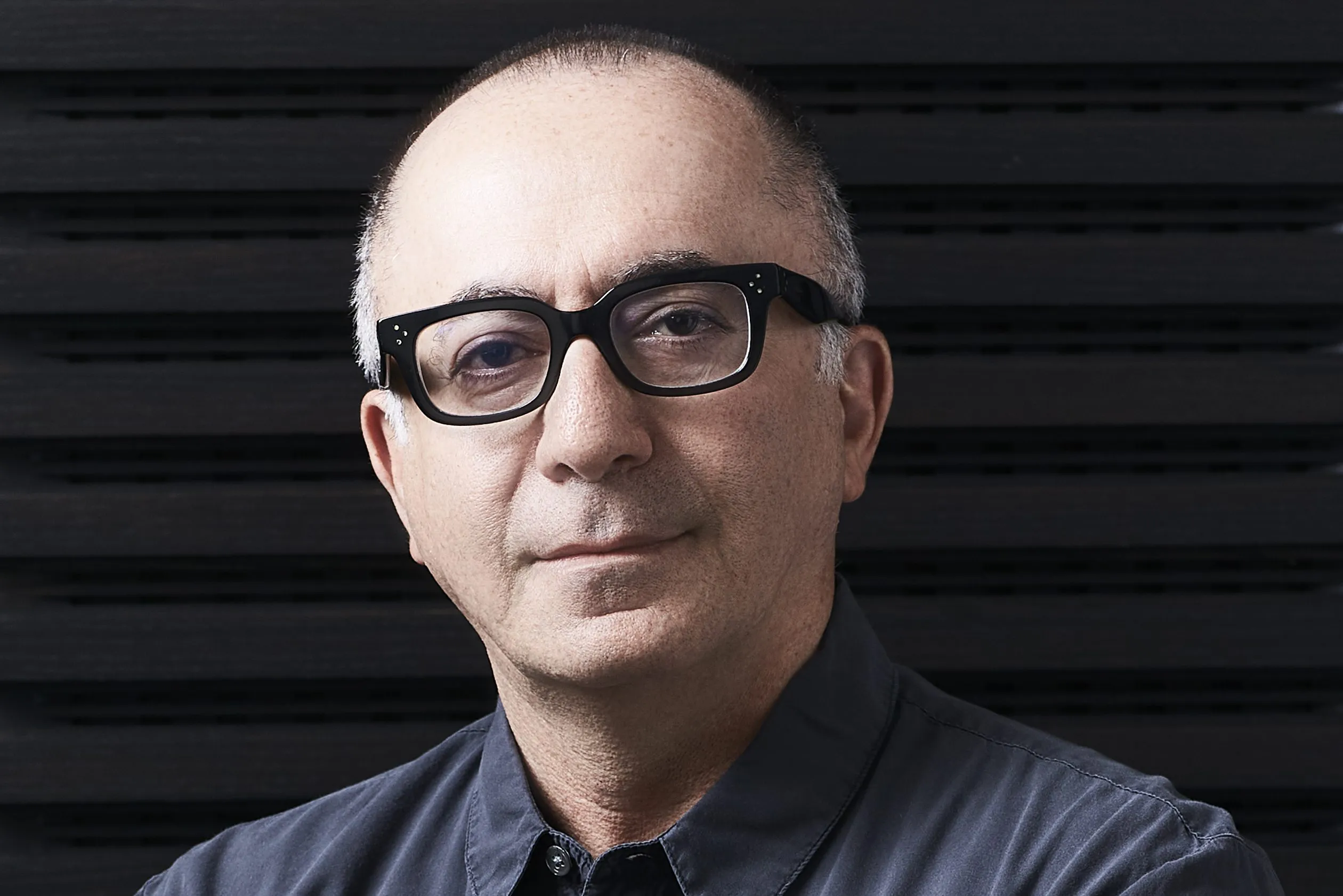Close-up portrait of a bald man wearing black glasses and a dark button-up shirt against a black slatted background.