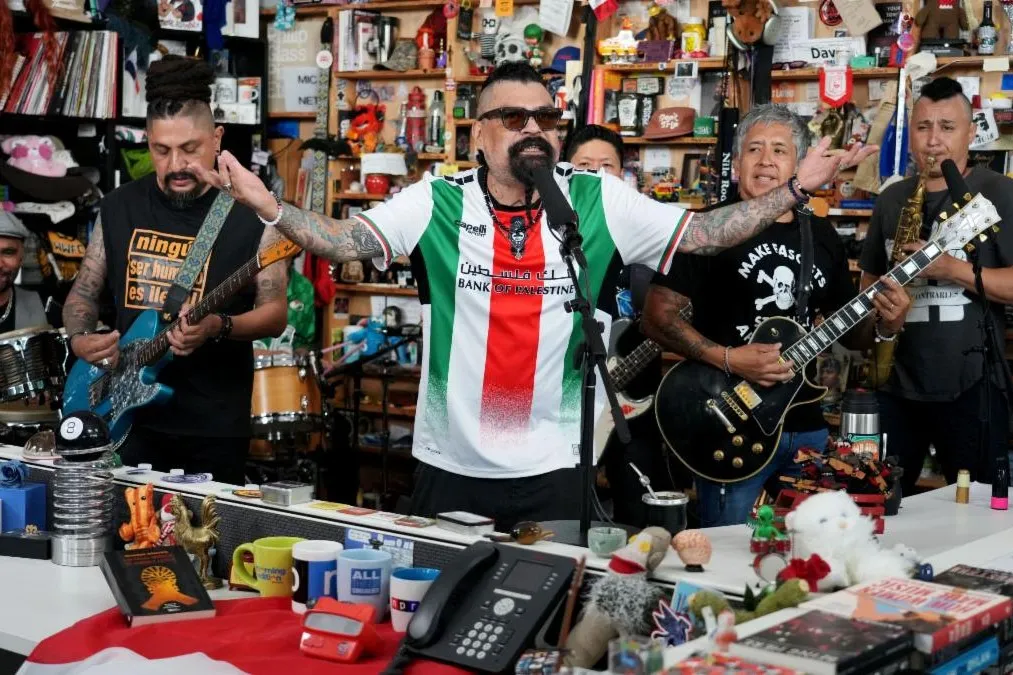 A singer with outstretched arms performs with guitarists and other musicians in a cluttered, colorful office.