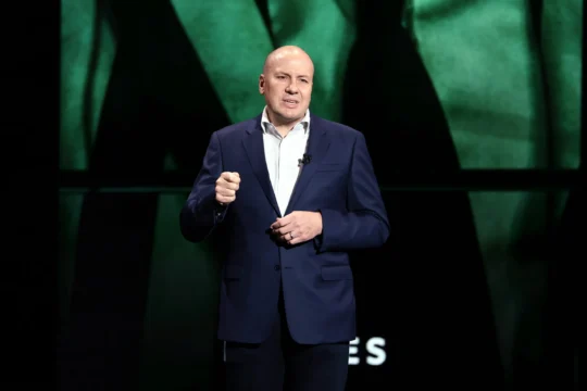 A bald man in a navy suit gestures while speaking onstage before a green abstract backdrop.