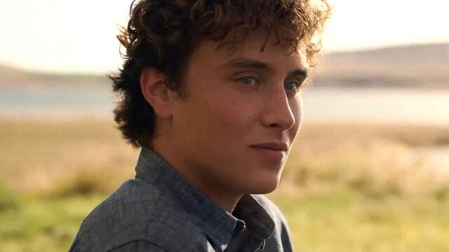A young man with curly hair in a blue shirt looks off to the side outdoors.