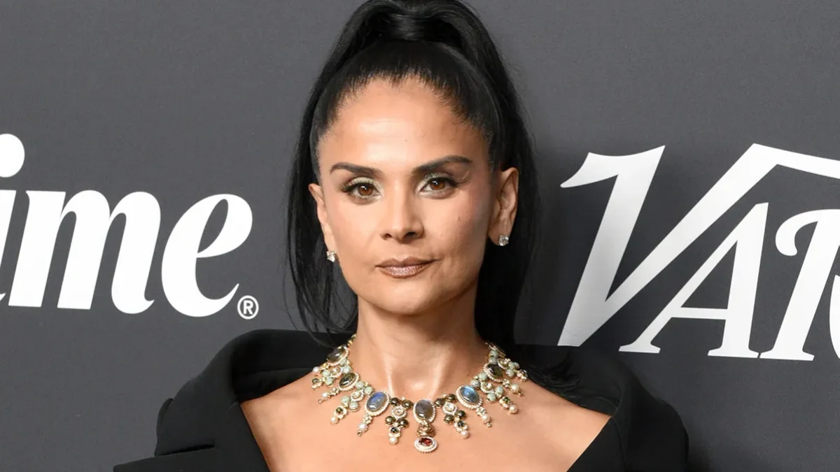 Woman with a high ponytail wears a black outfit and ornate necklace against a dark logo backdrop.