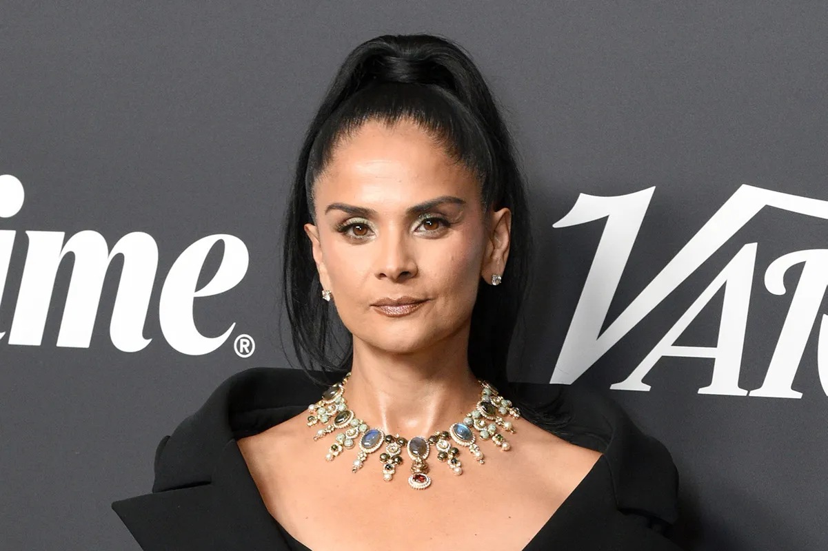 Woman with a high ponytail wears a black outfit and ornate necklace against a dark logo backdrop.