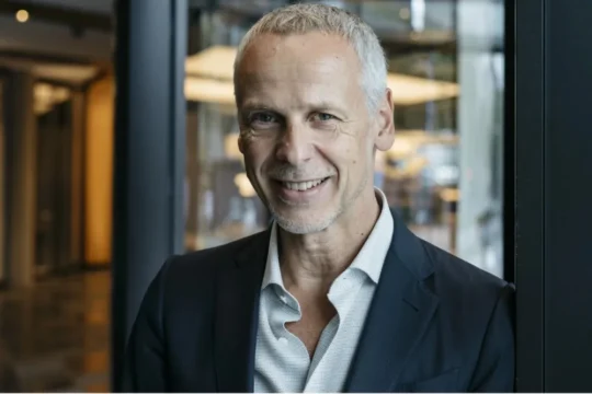 Smiling gray-haired man in a dark blazer stands in front of glass windows.