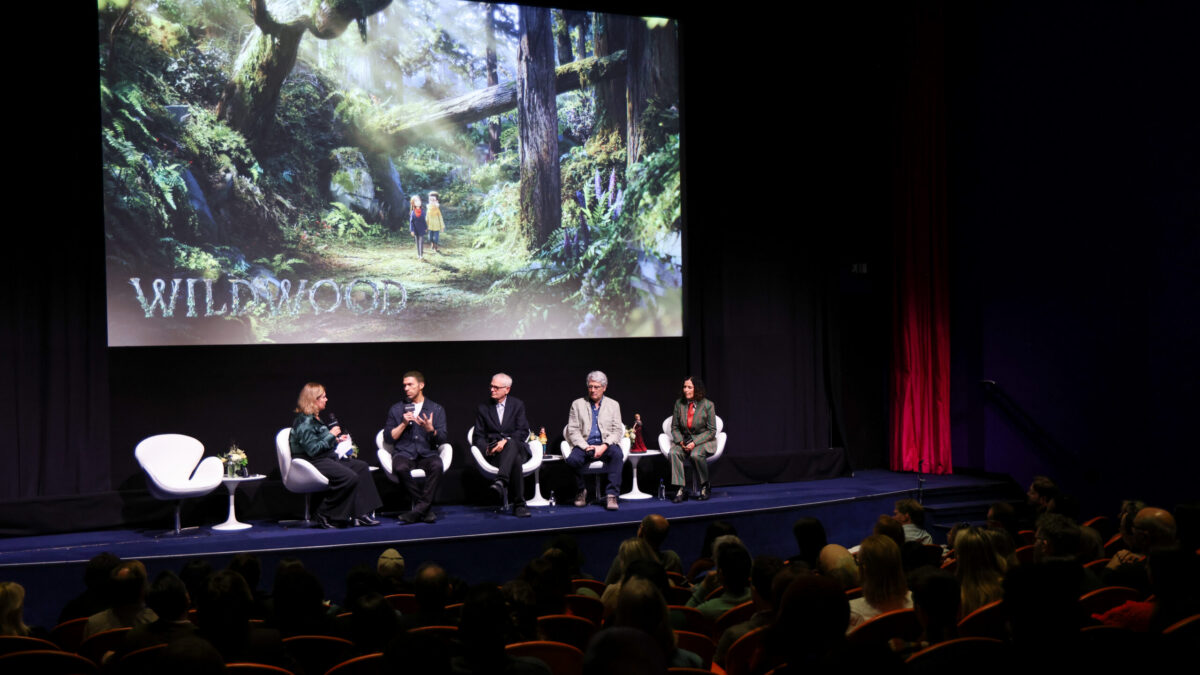 Five panelists sit onstage before an audience beneath a large forest scene projection reading “Wildwood.”