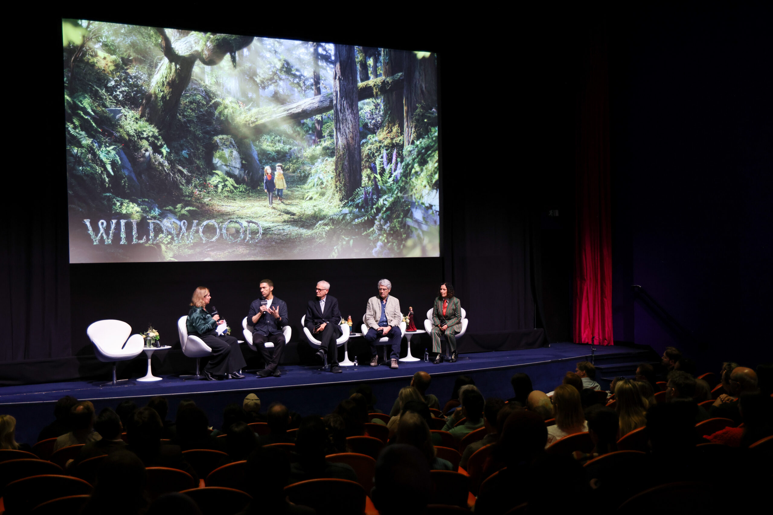 Five panelists sit onstage before an audience beneath a large forest scene projection reading “Wildwood.”