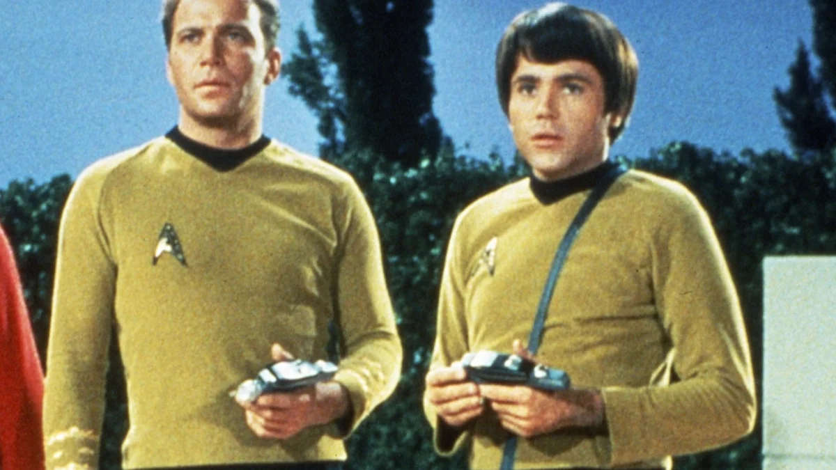 Two men in yellow futuristic uniforms hold small devices while standing outdoors against trees and sky.