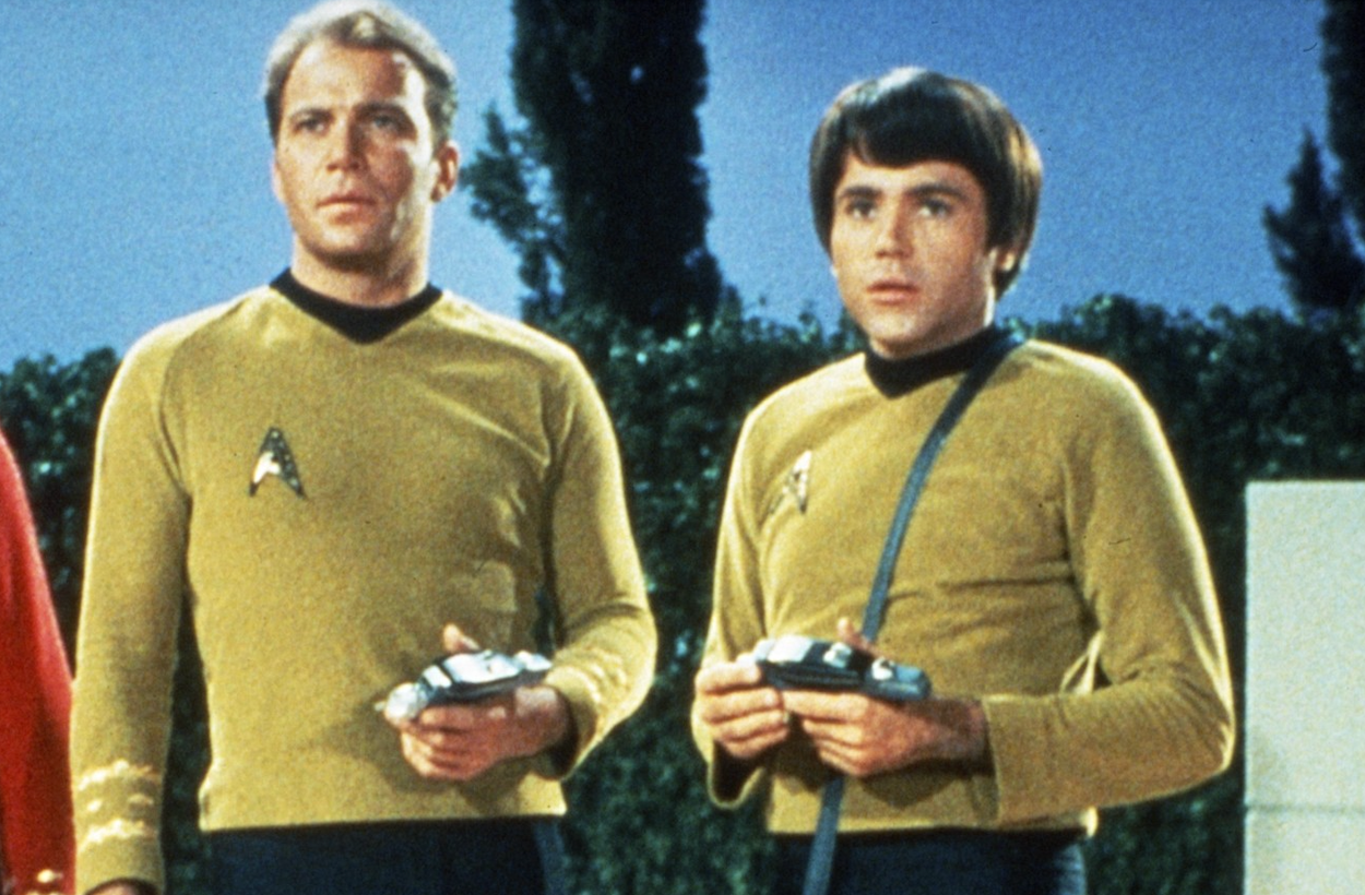 Two men in yellow futuristic uniforms hold small devices while standing outdoors against trees and sky.