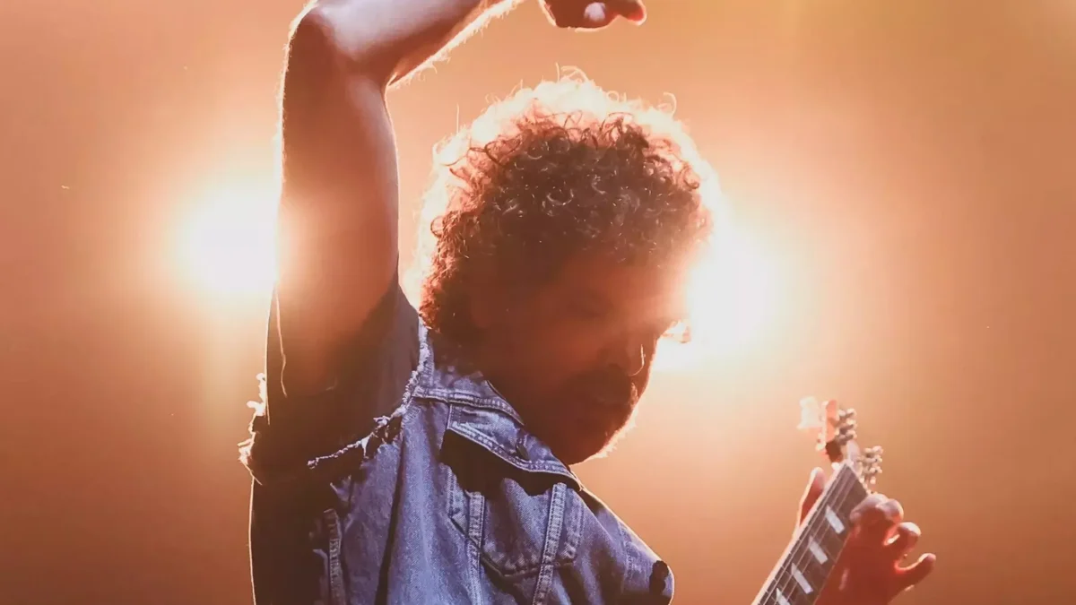 A curly-haired person raises one arm while holding a guitar under bright orange stage lights.