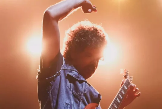 A curly-haired person raises one arm while holding a guitar under bright orange stage lights.